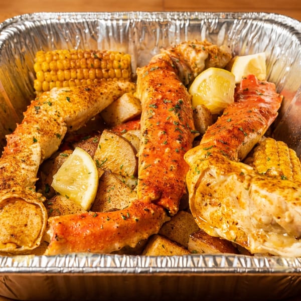 Seasoned king crab legs, corn, potatoes, and lemon wedges in a foil takeout pan.